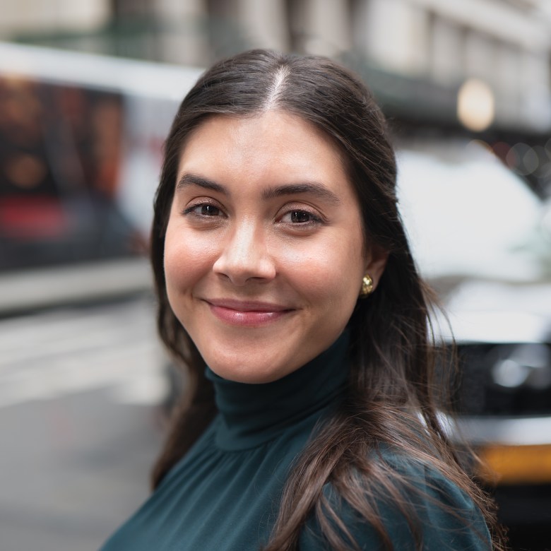 A woman soft smiles at the camera. She has brown hair and brown eyes and is wearing a green turtleneck.